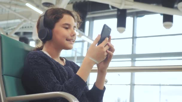 airport waiting hall room for a flight by plane. lifestyle young happy teen girl in headphones listening to the music on smartphone chatting communicates in the messenger. teenager girl in social