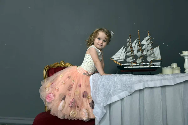 迷人的小女孩在优雅的白色与粉红色的礼服坐在一张桌子上与玩具帆船 复古风格的照片 — 图库照片