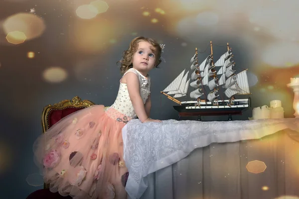 迷人的小女孩在优雅的白色与粉红色的礼服坐在一张桌子上与玩具帆船 复古风格的照片 — 图库照片