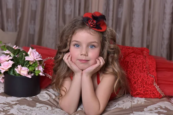 バラの箱とソファの上に座っている赤いドレスと帽子の小さな女の子王女の肖像 — ストック写真