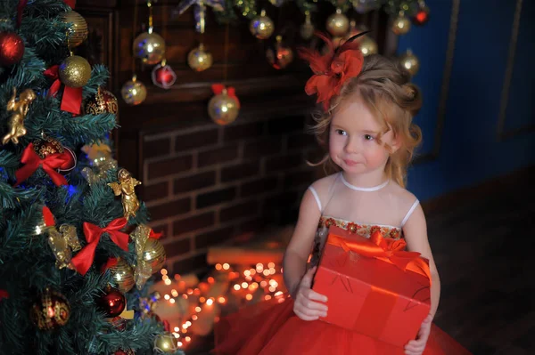 可爱的小女孩在圣诞树上的红色优雅的礼服与礼物在她的手中的盒子 — 图库照片