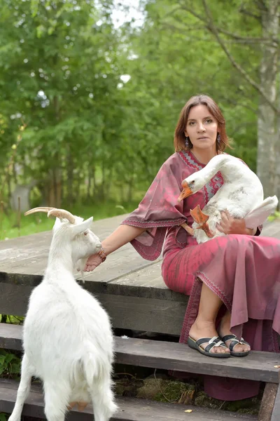 Woman in a burgundy dress on a farm with a goose in her arms and — Stock Photo, Image