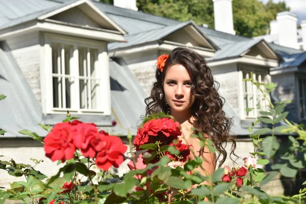 Schönes Mädchen mit Locken neben roten Rosen im Garten — Stockfoto