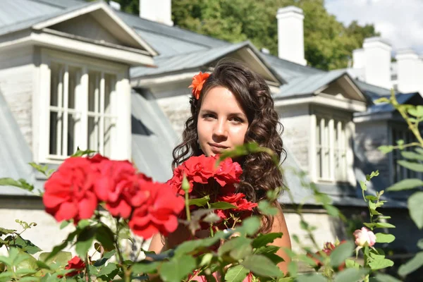 美丽的女孩与卷曲旁边的红玫瑰在花园里 — 图库照片
