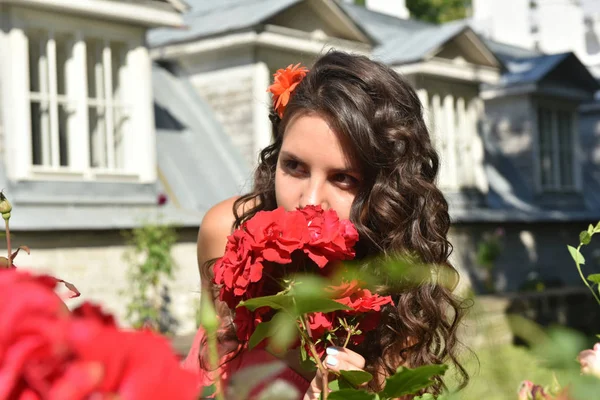 Beautiful girl with curls next to red roses in the garden — Stock Photo, Image