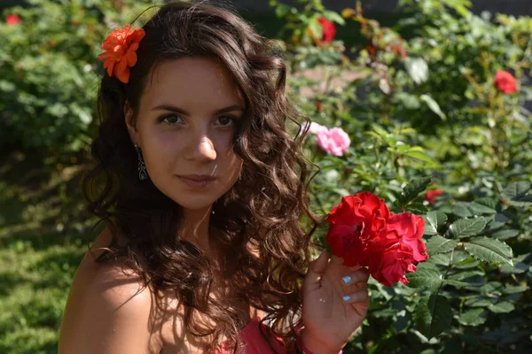 Schönes Mädchen mit Locken neben roten Rosen im Garten — Stockfoto