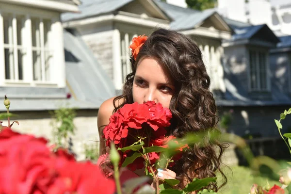 Krásná dívka s kadeřemi vedle červených růží v zahradě — Stock fotografie