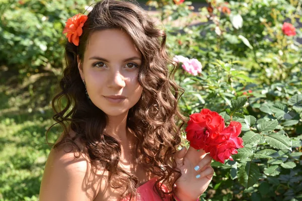 Beautiful girl with curls next to red roses in the garden — Stock Photo, Image