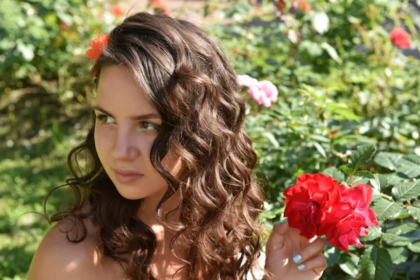Menina bonita com cachos ao lado de rosas vermelhas no jardim — Fotografia de Stock