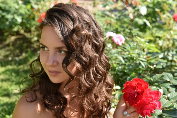 Beautiful girl with curls next to red roses in the garden — Stock Photo, Image