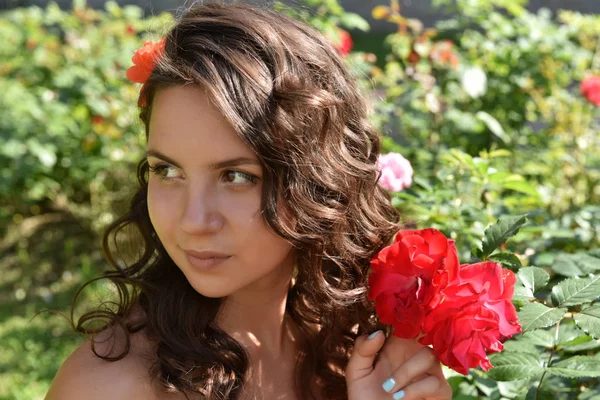 Красивая девушка с кудряшками рядом с красными розами в саду — стоковое фото