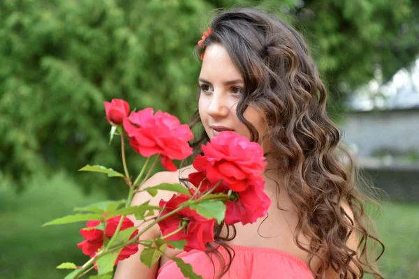 Piękna dziewczyna z loki obok czerwonych róż w ogrodzie — Zdjęcie stockowe