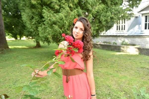 Beautiful girl with curls next to red roses in the garden — Stock Photo, Image