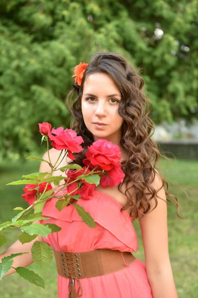 Красивая девушка с кудряшками рядом с красными розами в саду — стоковое фото