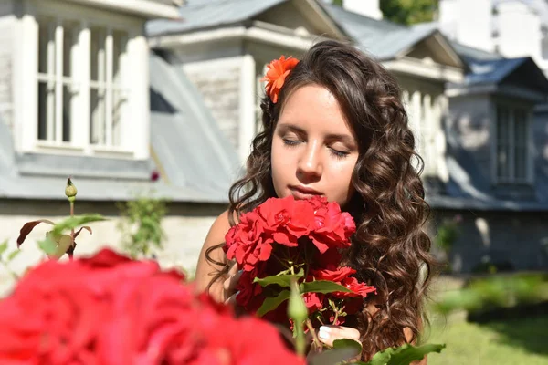 Beautiful girl with curls next to red roses in the garden — Stock Photo, Image