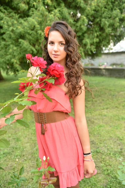 Beautiful girl with curls next to red roses in the garden — Stock Photo, Image