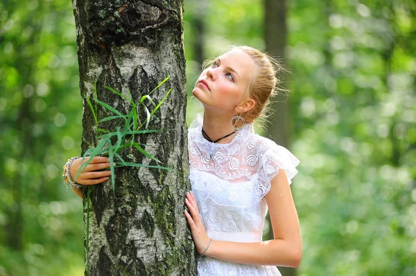 Blonde in een witte jurk staat in het park door de boom — Stockfoto