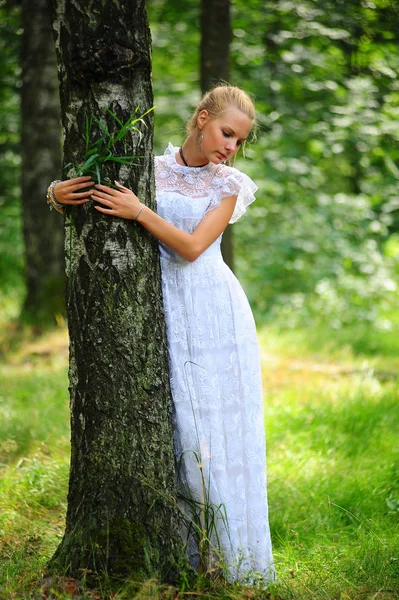 Ξανθιά σε ένα λευκό φόρεμα στέκεται στο πάρκο δίπλα στο δέντρο — Φωτογραφία Αρχείου