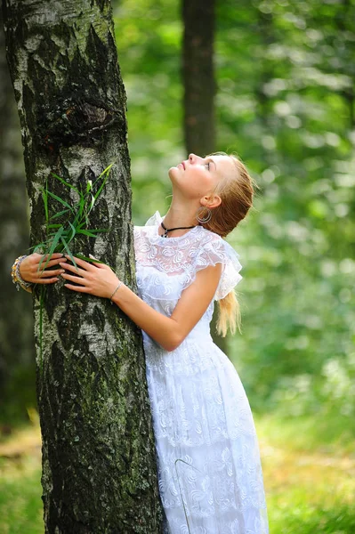 Blondýnka v bílých šatech stojí v parku u stromu — Stock fotografie