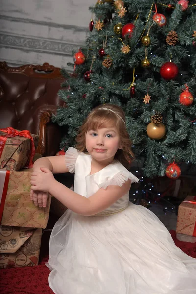 Menina em vestido branco sentado na árvore de Natal com presentes — Fotografia de Stock