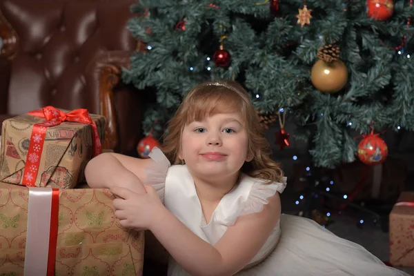 Menina em vestido branco sentado na árvore de Natal com presentes — Fotografia de Stock