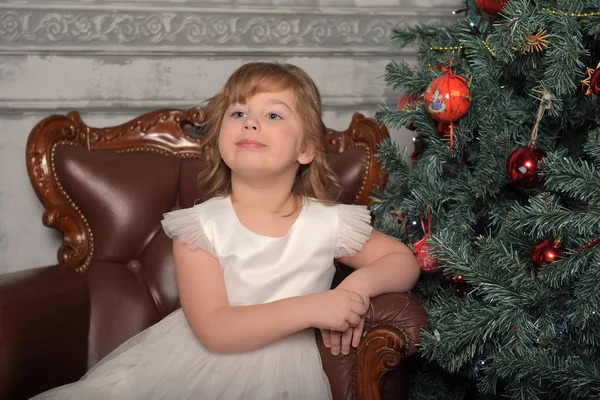 ドレスを着た女の子はクリスマスツリーの前の古い椅子に座っている — ストック写真