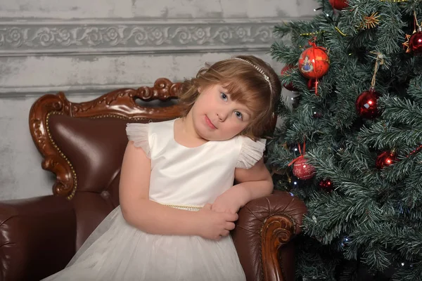 Menina de vestido senta-se em uma cadeira velha pela árvore de Natal — Fotografia de Stock