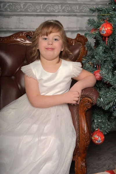 Chica en vestido se sienta en una silla vieja junto al árbol de Navidad —  Fotos de Stock