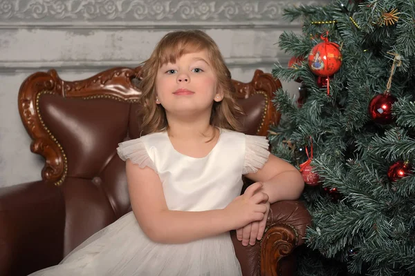 Menina de vestido senta-se em uma cadeira velha pela árvore de Natal — Fotografia de Stock