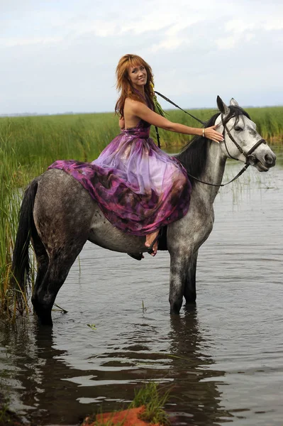 Donna in un abito lungo viola cavalcando un cavallo in acqua in — Foto Stock