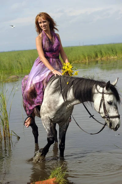 Γυναίκα σε ένα μωβ μακρύ φόρεμα ιππασία ένα άλογο στο νερό στο — Φωτογραφία Αρχείου