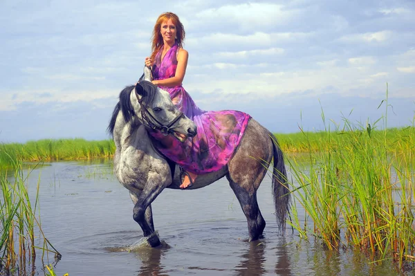 Žena ve fialových dlouhých šatech na koni ve vodě — Stock fotografie