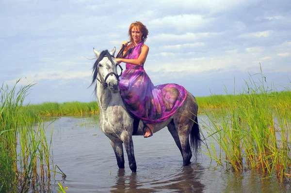 Mulher em um vestido longo roxo montando um cavalo na água no — Fotografia de Stock