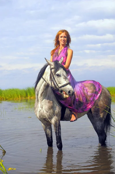 穿着紫色长裙的女人在水里骑马 — 图库照片