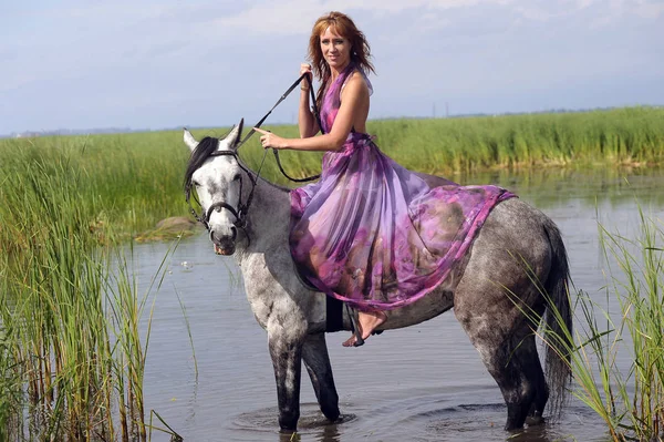 Mujer en un vestido largo púrpura montar a caballo en el agua en el — Foto de Stock