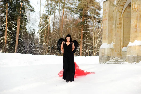 Mädchen mit schwarzen Engelsflügeln in einem schwarzen Kleid im Winter und roten — Stockfoto