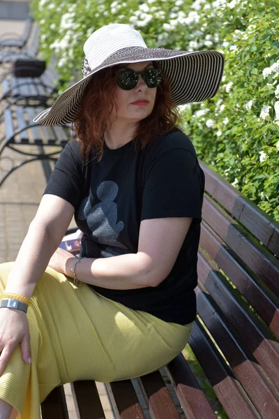 Uma mulher de chapéu e óculos de sol está sentada em um banco — Fotografia de Stock