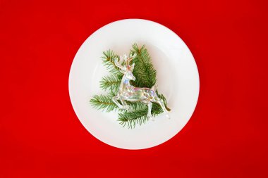 Noel ve Yeni Yıl için şenlik masası. Boş beyaz tabak, şenlikli süslemeler kürk ağacı dalları ve kırmızı arka planda geyik. Tatil süsleri. Üst görünüm. 