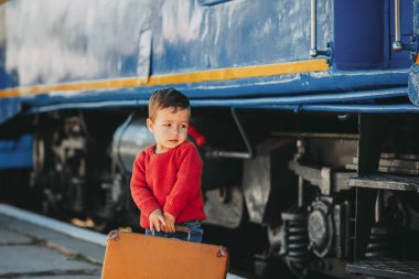 Sevimli küçük çocuk çocuk bir tren istasyonu retro eski kahverengi çanta trenin yanında kırmızı kazak giymiş. Tatil için hazır. Platformda genç yolcusu.