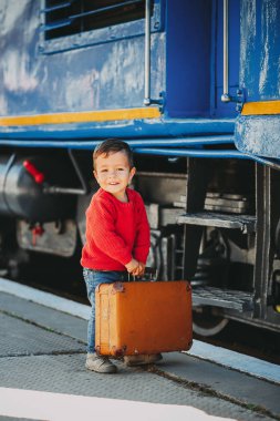 Sevimli küçük çocuk çocuk bir tren istasyonu retro eski kahverengi çanta trenin yanında kırmızı kazak giymiş. Tatil için hazır. Platformda genç yolcusu.