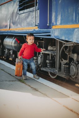 Sevimli küçük çocuk çocuk bir tren istasyonu retro eski kahverengi çanta trenin yanında kırmızı kazak giymiş. Tatil için hazır. Platformda genç yolcusu.