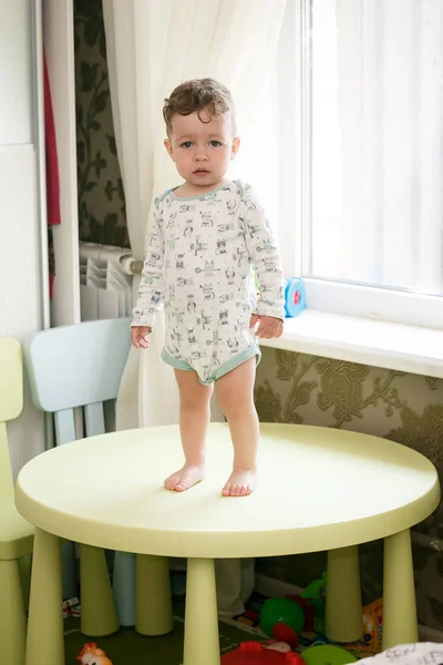 The baby curly-headed boy is standing on the table on window bac — Stock Photo, Image