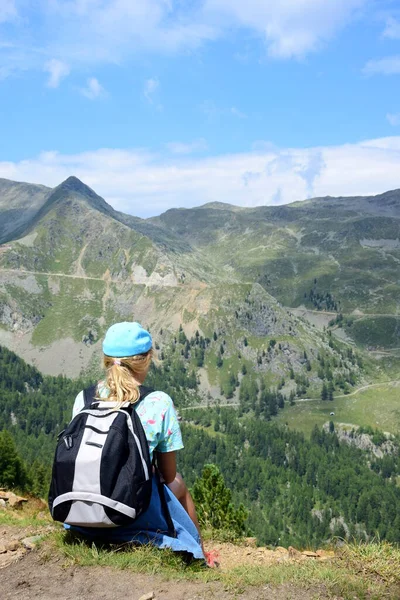 Young girl with hiking clothes and a backpack sits on a hill while hiking and looks at the surrounding mountains in the Ultental in South Tyrol, Italy