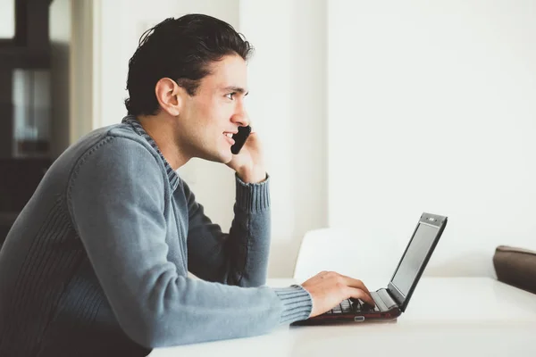 Schöner Junger Mann Sitzt Drinnen Und Spricht Smartphone Mit Computer — Stockfoto