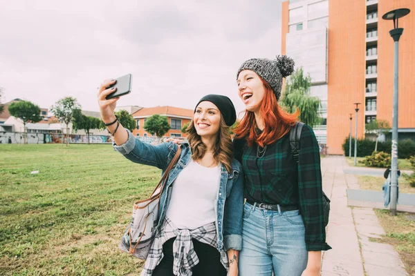 两个年轻的妇女走在户外采取自拍使用智能手机 — 图库照片
