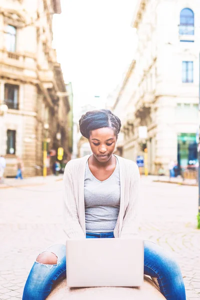 年轻非洲妇女使用计算机户外在城市坐在长凳敲击键盘 — 图库照片