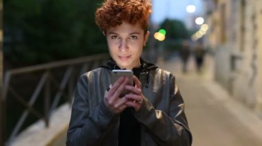 Beyaz saçlı genç, kıvırcık saçlı kadın gün batımında elinde ekranın ışığıyla aydınlanan akıllı bir telefonla kameraya bakıp gülüyor.