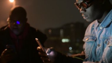 4k. Genç siyahi adam gece açık havada akıllı telefon ile aydınlatılmış ekran ışığı kablosuz internet teknolojisi, sosyal ağ, kablosuz iletişim konsepti kullanarak
