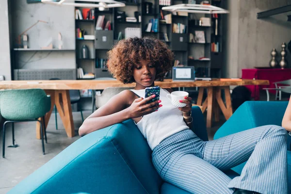 Ung svart kvinna dricka och chatta med smartphone i moderna loft — Stockfoto