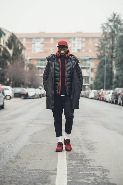 Молодой бородатый африканский мужчина на открытом воздухе ходит, глядя в камеру улыбается
